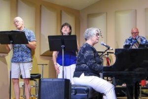 Two choir mambers standing and sining while Julie Keefer plays the piano